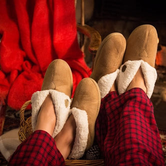A pair of beige colored faux suede ladies' cozy slippers with faux sheepskin lining, worn with red plaid pajama pants.