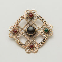 An ornate Celtic brooch featuring a cross design with interwoven patterns. It has four decorative squares, each containing a small gemstone: two red and one green, with a central black stone. The piece combines intricate metalwork with colored accents.