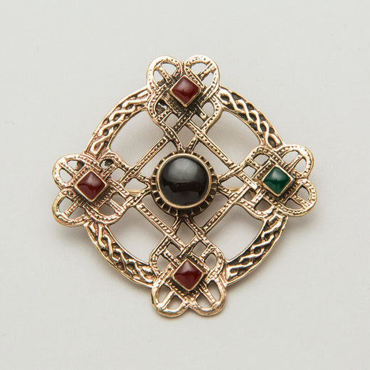 An ornate Celtic brooch featuring a cross design with interwoven patterns. It has four decorative squares, each containing a small gemstone: two red and one green, with a central black stone. The piece combines intricate metalwork with colored accents.