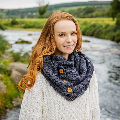 A woman standing by a river, wearing a gray wool snood with wooden buttons over a white knit sweater.