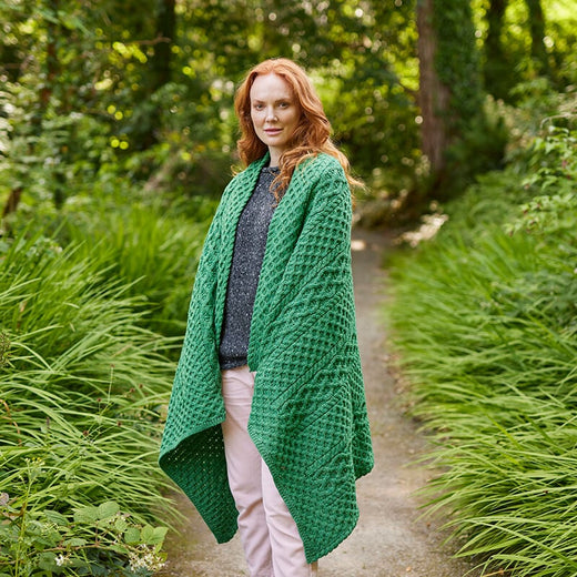 A woman with long red hair stands on a path surrounded by lush green grass and foliage. She is wearing a gray sweater and pink pants, wrapped in a textured green Aran blanket. The natural setting provides a serene atmosphere.