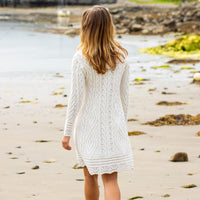 A woman with long blonde hair walks along a sandy beach, wearing a cozy white knitted Aran dress. The shoreline is visible in the background, with rocks and patches of greenery nearby.