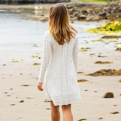 A woman with long blonde hair walks along a sandy beach, wearing a cozy white knitted Aran dress. The shoreline is visible in the background, with rocks and patches of greenery nearby.