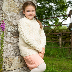 A young girl leans against a stone wall, wearing a cream-colored kids Aran sweater and a pale pink skirt. She has long hair parted to the side and stands in a grassy area with greenery and a gate visible in the background.