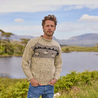A man standing outdoors wearing a beige Aran sweater with a sheep landscape design.
