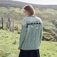 Image featuring the Sheep Aran Sweater in green, crafted with authentic Irish Aran cable knit patterns and a unique sheep and mountain-inspired design across the back, showcasing traditional Irish craftsmanship in a rugged rural setting.