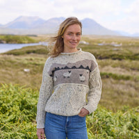 A woman standing outdoors wearing a beige Aran sheep sweater with a landscape design, made from pure wool.