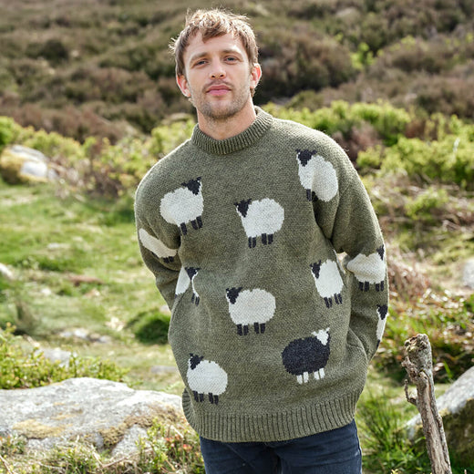 Man wearing a green sweater with sheep pattern standing in a natural landscape.