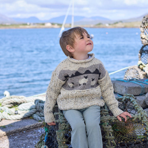 A young child sits on a fishing net near a body of water, wearing a cream-colored sweater with a pattern. The background features boats and distant hills under a partly cloudy sky. The child looks up thoughtfully, enjoying the outdoors.