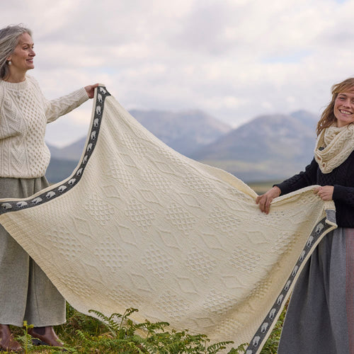 Two people holding up a cream Aran Sheep Throw outdoors, showing its textured knit pattern and decorative sheep‑themed border with mountains in the background.