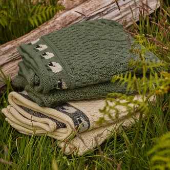 Two folded Aran wool blankets in green and beige, featuring a traditional geometric pattern, laid on grass beside a wooden log.