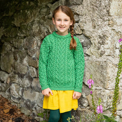 A young girl stands against a stone wall, smiling at the camera. She wears a green kids Aran sweater and a bright yellow skirt, with her hair styled in a braid. Pink flowers grow nearby, adding color to the scene.