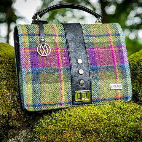A multicolored tweed handbag with a black leather handle and metal twist clasp, sitting on a mossy surface.