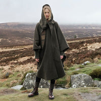 A model stands outdoors in a rugged landscape, wearing a dark green Irish walking cape over and brown leather boots. The background features rolling hills and rocky terrain under a cloudy sky.