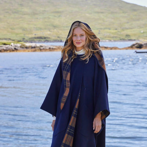 A woman stands by a body of water, wearing a dark blue cape with a tartan pattern. Her long hair cascades down as she smiles at the camera. The background features a green hillside and a clear sky.
