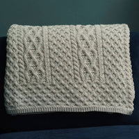 A textured, beige knitted Aran blanket drapes over a dark blue couch in a  The wall behind is painted a muted green, complementing the overall warm aesthetic.