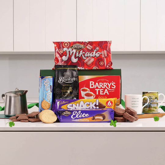 A snack and tea gift basket arranged on a kitchen counter with white cabinets behind it. Visible products include a red bag of Scotts Mikado candy, a green box of Bailey's Gold Mint, a red bag of Barry's Tea, a purple bag of Snack! Elite chocolate biscuits, a blue bag of Oreo, a black bag of Ben & Jerry's, a silver kettle, chocolate biscuits, a round biscuit, and two mugs.