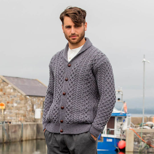 A man wearing a gray Aran cardigan with a high shawl collar and traditional brown leather buttons, standing in front of a harbor scene.