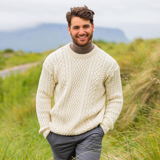 A man standing in a field wearing a classic Aran sweater in a cream color.