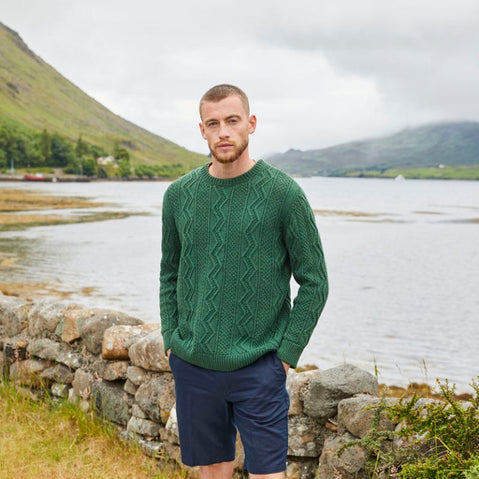 A man wearing a green crew neck aran sweater featuring a traditional zig-zag stitch design, standing by a stone wall with a scenic view of a lake and mountains in the background.