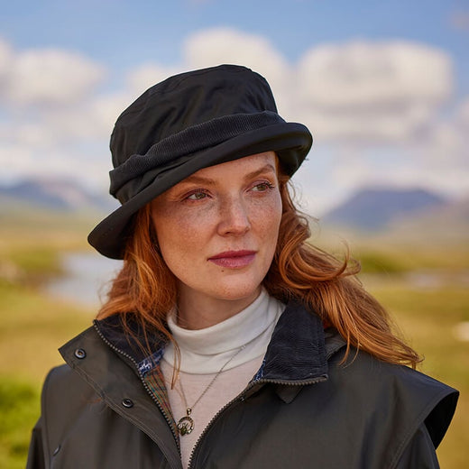 A woman with curly red hair wears a black bucket hat and a dark jacket, standing outdoors against a landscape of rolling hills and a cloudy sky. She looks thoughtfully into the distance, with greenery surrounding her.
