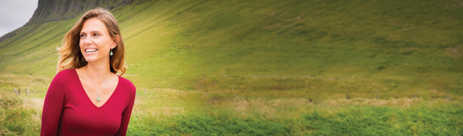 A woman in a red top and wearing celtic jewelry stands smiling against a backdrop of green rolling hills. The scene conveys a sense of warmth and nature.