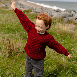 A child with red hair wears a cozy red kids Aran sweater, standing on grass near the shore. They have arms outstretched and are smiling, with rocky beach and ocean in the background.