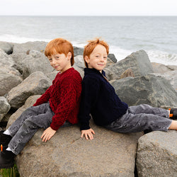 Two young boys with red hair sit on large rocks by the shore. One boy wears a red kids Aran sweater, while the other is in a navy blue sweater. They are positioned back-to-back, smiling at the camera, with a view of the ocean in the background.