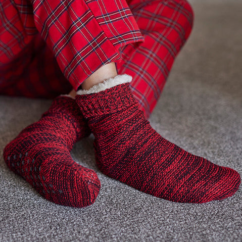 A pair of red and black fleece lined men's socks with a non-slip sole, worn on a person sitting cross-legged.