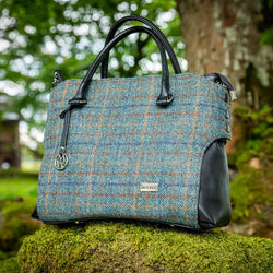 A handbag with a tweed body and vegan leather details, placed on a stone surface with foliage in the background.