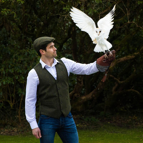 A person wearing a green Irish wool Trinity flat cap holding a white owl on their extended hand, outdoors with trees in the background.