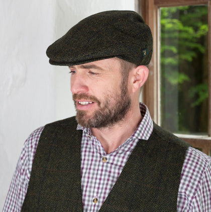 A man with a beard is wearing a dark flat cap and a checked shirt underneath a dark vest. He is standing near a window, with greenery visible outside. The focus is on his profile as he looks slightly to the side.