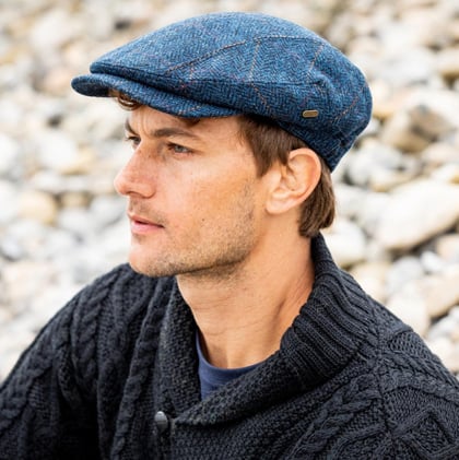 A man with short hair sits on a stony surface, wearing a blue tweed flat cap and a dark, knitted sweater. He gazes thoughtfully to the side, with soft natural lighting illuminating his features.