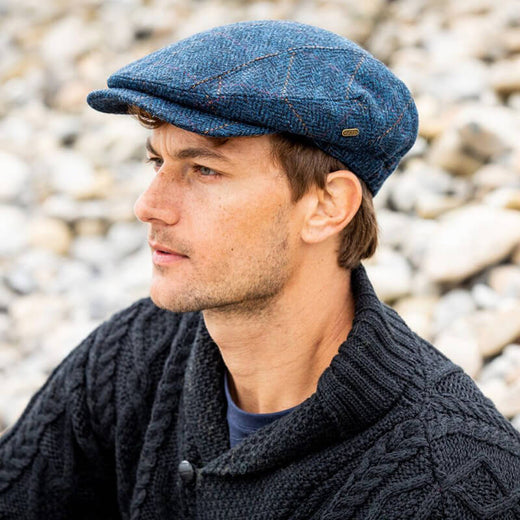 A man with short brown hair sits on a rocky surface, wearing a navy blue Irish tweed flat cap and a dark cable-knit sweater. He appears contemplative, looking off into the distance.