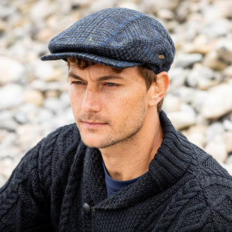 A man with short brown hair sits on a rocky surface, wearing a navy blue Irish tweed flat cap and a dark cable-knit sweater. He appears contemplative, looking off into the distance.
