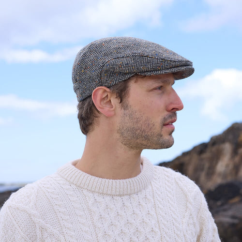 A man wearing a gray Irish flat cap with a herringbone pattern, in a natural outdoor setting.