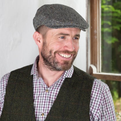 A smiling man with a beard stands in front of a window, wearing a gray flat cap and a green vest over a checkered shirt. Natural light illuminates his face, highlighting the casual yet stylish outfit.