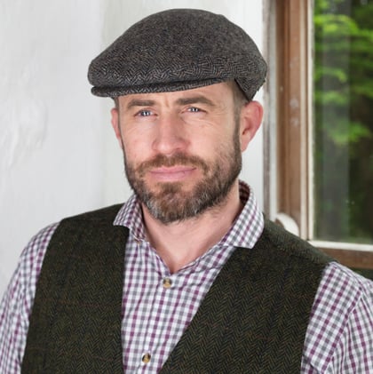 A man with a beard and a serious expression stands in front of a window. He wears a gray flat cap, a checkered shirt, and a dark vest. Soft natural light illuminates the scene, highlighting the textures of his clothing and the interior environment.