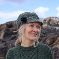 A woman with long, light-colored hair smiles while wearing a green sweater and a plaid cap. She stands outdoors by rocky terrain under a cloudy sky.