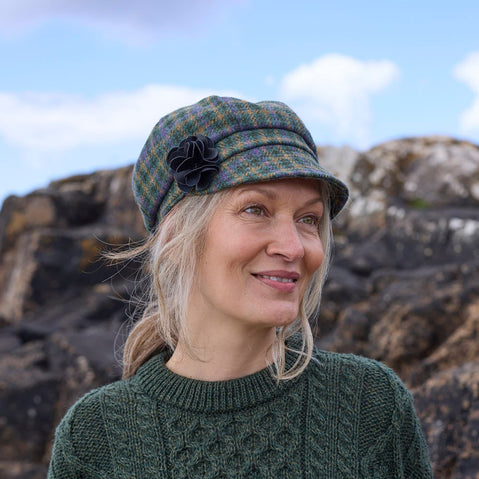 A woman with long, light-colored hair smiles while wearing a green sweater and a plaid cap. She stands outdoors by rocky terrain under a cloudy sky.