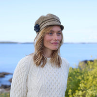 A woman stands by a body of water, wearing a cream-colored cable-knit sweater and a light brown hat adorned with a flower. Her hair is slightly wavy, and she gazes softly into the distance. In the background, the blue water meets a clear sky, while yellow flowers are visible nearby.