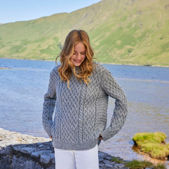 A woman standing by a lake wearing a gray Aran sweater with a ribbed turtle neck and cuffs.
