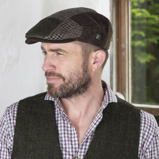 A man with a beard wearing an Irish tweed flat cap and a checked shirt under a dark vest stands near a window, looking thoughtfully to the side. Natural light illuminates the scene, highlighting the indoor setting.