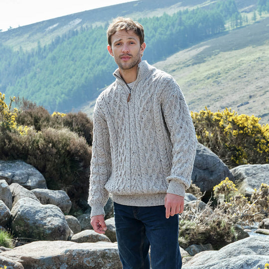 Man wearing a cable knit sweater standing in a mountainous landscape with rocks and greenery.