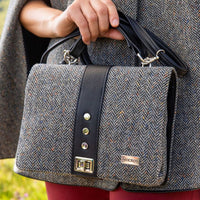 Fiona tweed handbag with black leather trim and handles, featuring a herringbone pattern and metal clasp, held at the side of a person wearing a matching tweed garment.