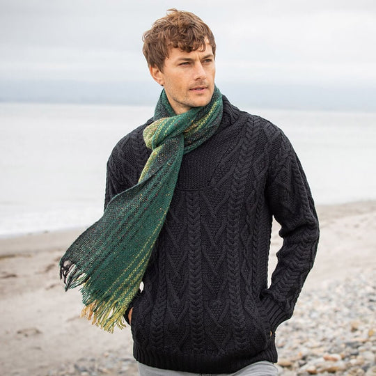 A man standing by the sea wearing a black knit sweater and a green alpaca wool scarf with a geometric pattern.