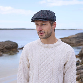 A man stands on a rocky beach, wearing a cable-knit white sweater and a dark flat cap. He gazes thoughtfully into the distance, with calm water and a cloudy sky in the background.