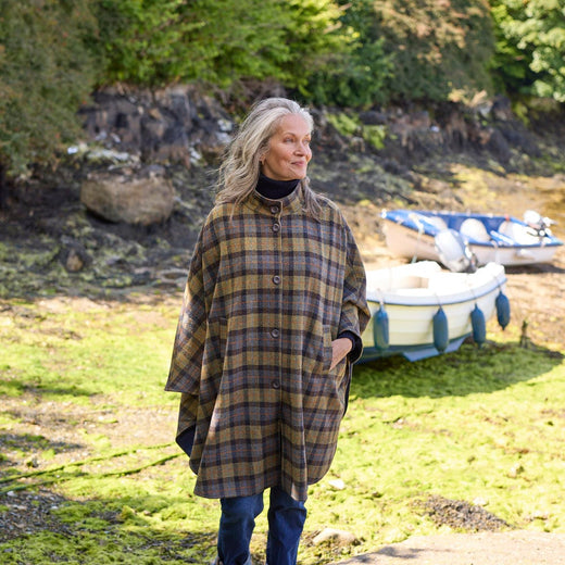 A woman with long, wavy hair stands on a rocky shoreline wearing a plaid, oversized cape. She gazes thoughtfully towards a small boat moored nearby, surrounded by greenery and water.
