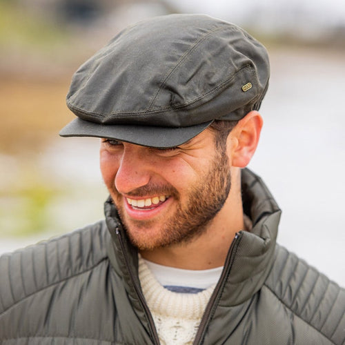 Olive waxed Irish flat cap with subtle panel stitching and a small brass logo tag, worn outdoors with a quilted jacket and cream sweater.