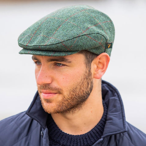 A man with a short beard is wearing a green Irish tweed flat cap and a navy jacket. He is looking slightly to the side, against a neutral background.